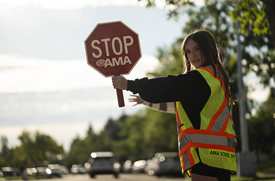 A school patroller with long brown hair and wearing a school patrol safety vest holds out her hand-held stop sign at a crosswalk.